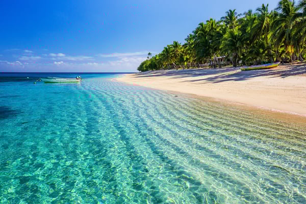 Türkises Meer mit kleinem Boot vor weissem Sandstrand und Palmen unter blauem Himmel