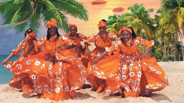 Gruppe von Frauen in leuchtend orangefarbenen traditionellen Kleidern tanzt fröhlich am Strand vor Palmen und einem Sonnenuntergang über dem Meer