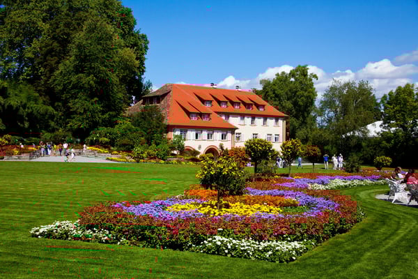 Jardins fleuris de l’île de Mainau avec parterres colorés, pelouse soignée et bâtiment historique en arrière-plan.