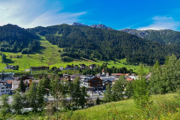 Village alpin de St. Anton am Arlberg, entouré de prairies vertes, de forêts et de montagnes sous un ciel bleu.