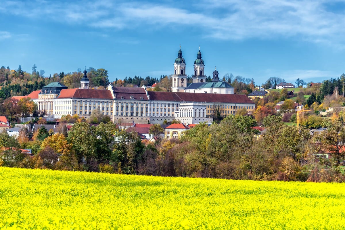 Abbaye baroque de Saint-Florian vue de loin, avec ses tours jumelles dominant un paysage automnal et un champ jaune au premier plan.