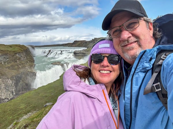 Couple souriant devant une grande cascade islandaise, paysage naturel sauvage en arrière-plan.
