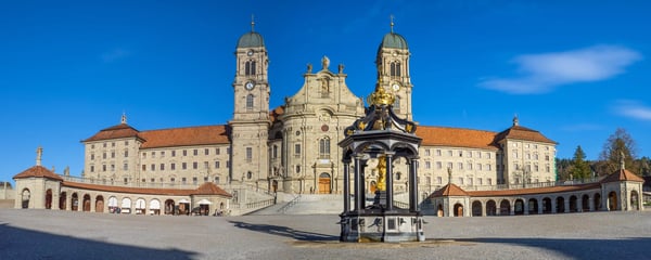 Abbaye baroque d’Einsiedeln avec façade monumentale et fontaine sur une vaste place.