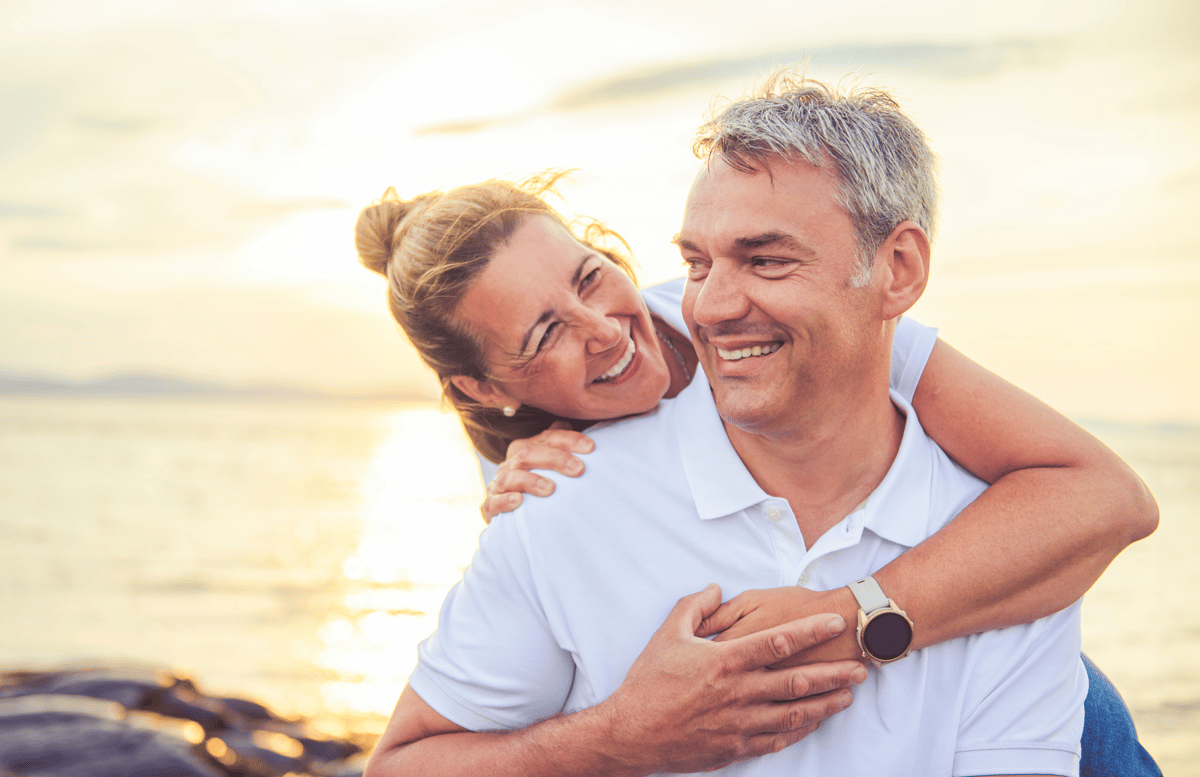 Couple souriant enlacé au bord de la mer au coucher du soleil.