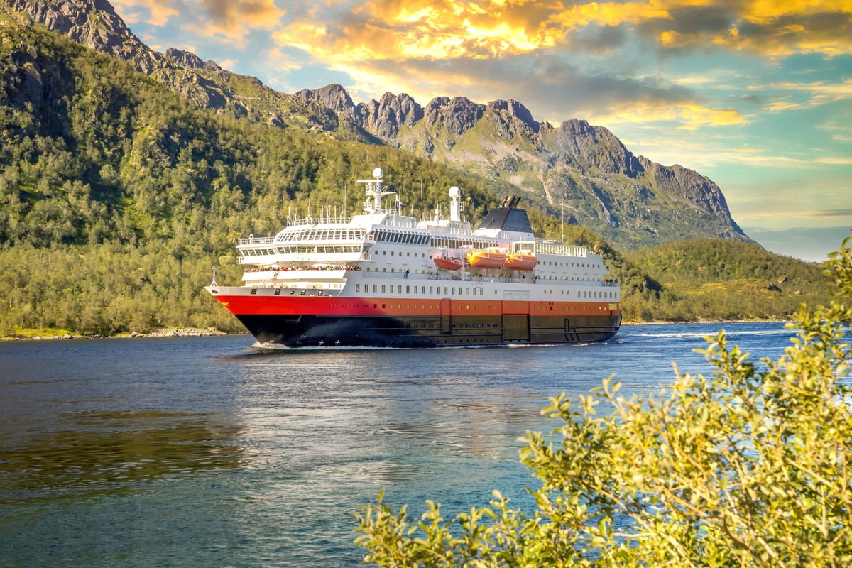 Hurtigruten-Schiff fährt durch einen Fjord vor bewaldeten Bergen im Abendlicht.