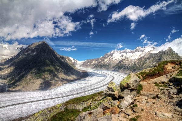 Vue panoramique du glacier d Aletsch serpentant entre des sommets rocheux, sentier de randonnée au premier plan et ciel partiellement nuageux