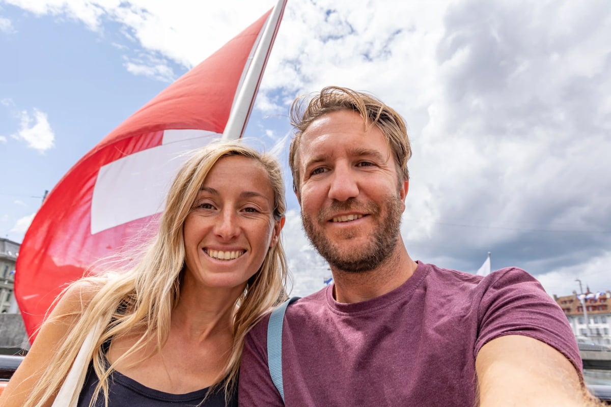 Couple souriant prenant un selfie en plein air devant un drapeau suisse rouge et blanc, ciel nuageux en arrière plan
