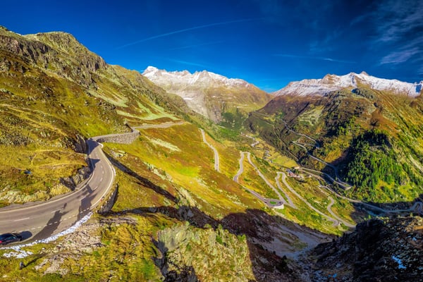 Route alpine sinueuse au col du Grimsel, traversant des montagnes verdoyantes avec sommets enneigés sous un ciel bleu.