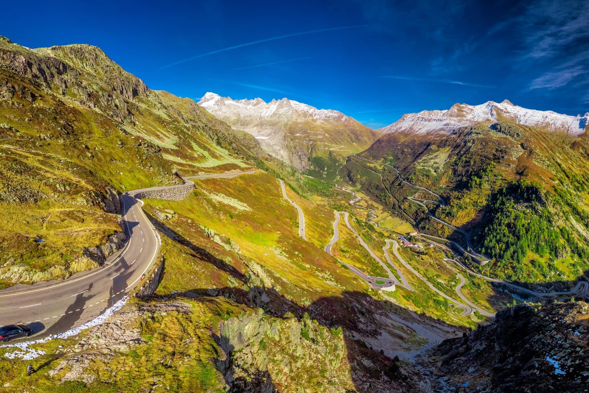 Route alpine sinueuse au col du Grimsel, traversant des montagnes verdoyantes avec sommets enneigés sous un ciel bleu.