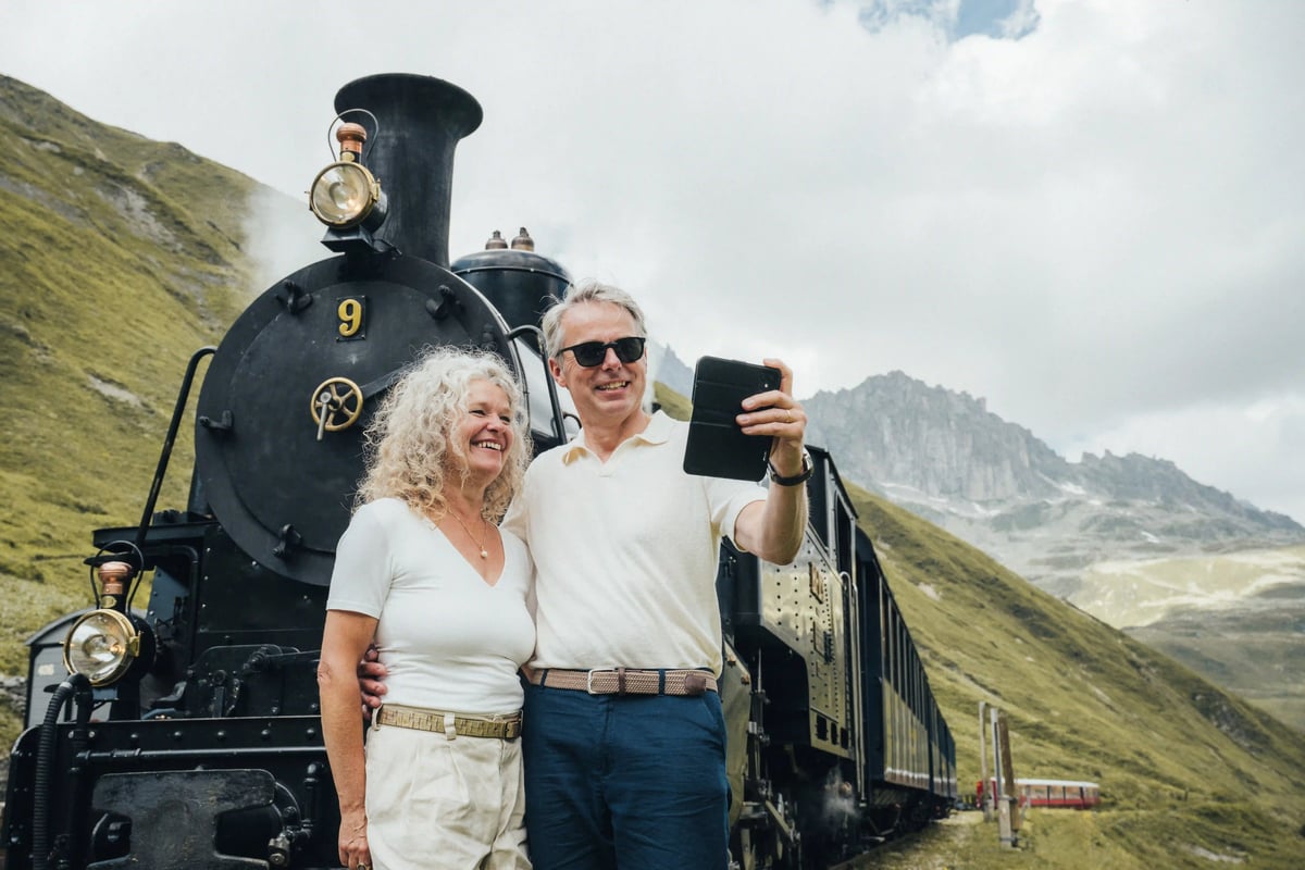 Couple souriant prenant un selfie devant une locomotive à vapeur historique de la ligne Furka, en paysage alpin.
