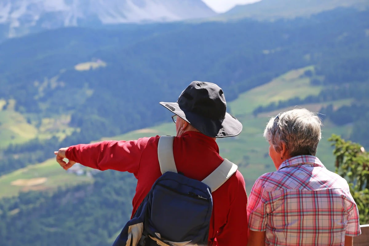 Deux randonneurs âgés regardent la vallée alpine depuis un point de vue, l’un d’eux montre le paysage de montagnes verdoyantes