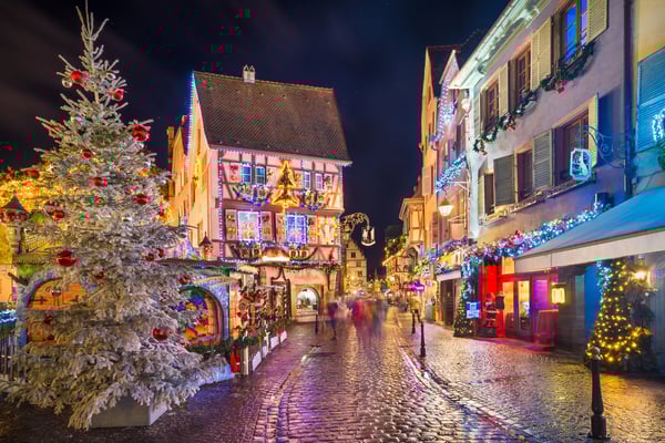 Rue de Colmar décorée pour Noël avec maisons à colombages illuminées, guirlandes colorées et sapin enneigé au premier plan