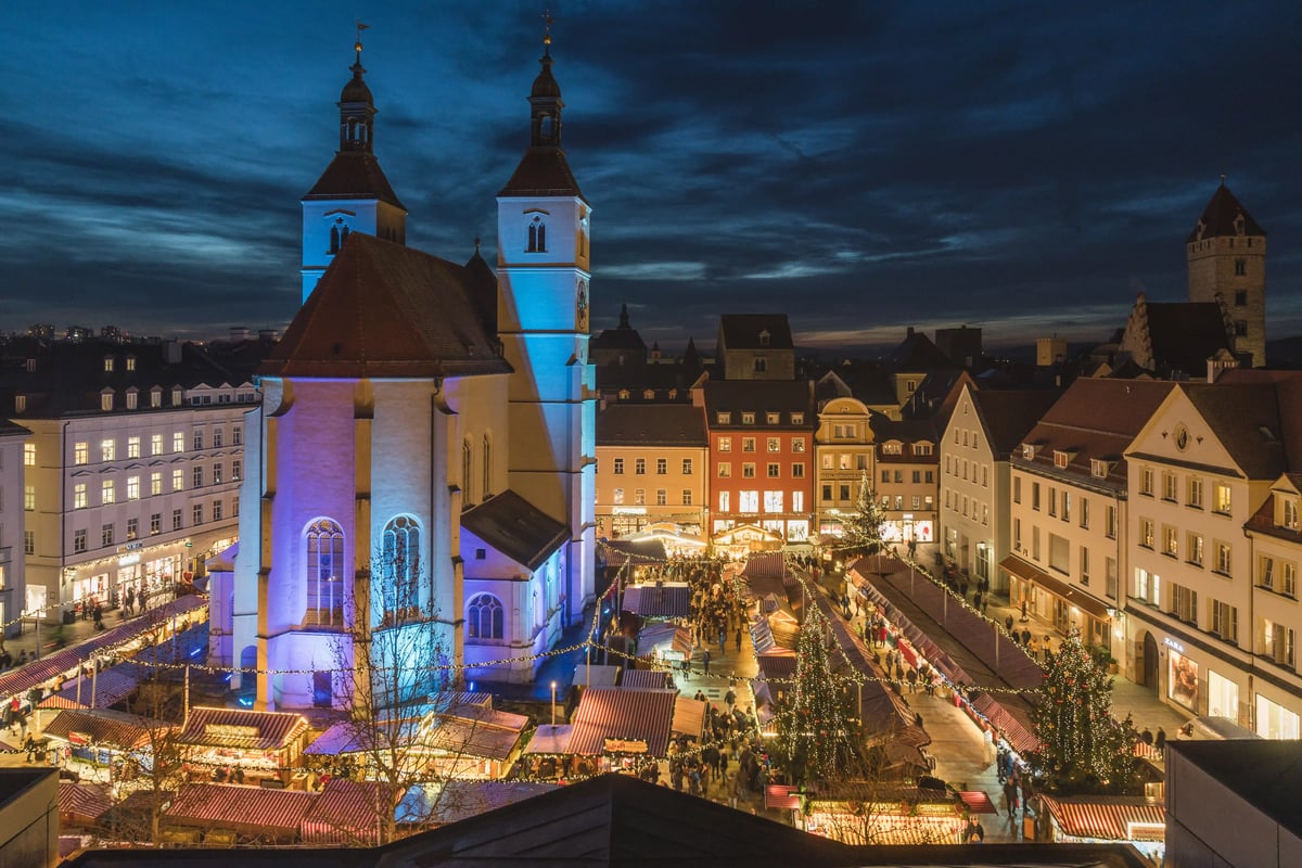 Abendlicher Weihnachtsmarkt in Regensburg mit beleuchteter Kirche, Marktstanden und Besuchern auf dem Platz.
