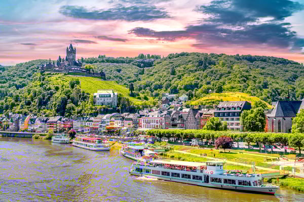 Flussufer mit Ausflugsschiffen vor Cochem, darüber Burg auf bewaldetem Hügel bei farbigem Abendhimmel.