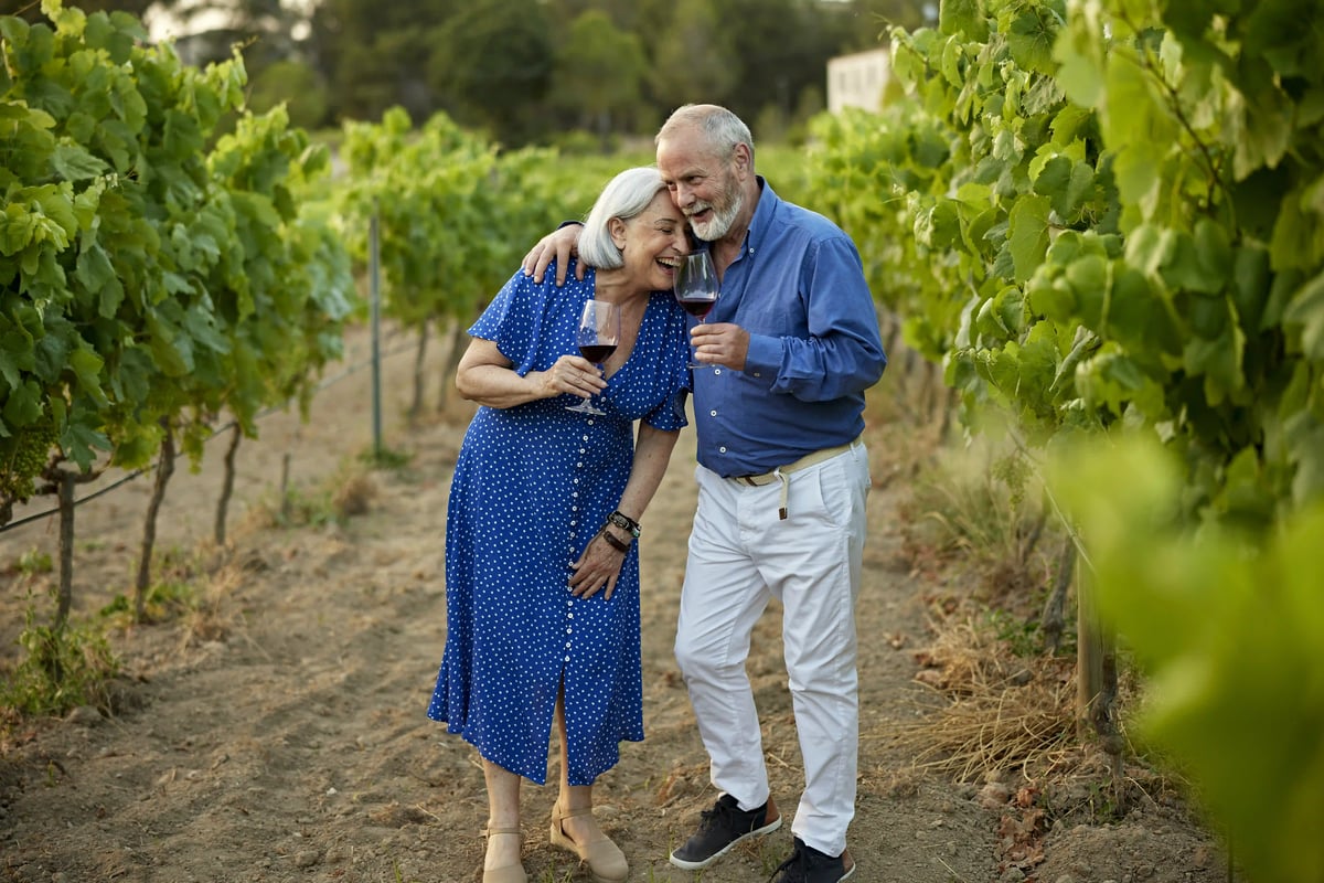 Älteres Paar lacht im Weinberg und hält zwei Gläser Rotwein zwischen grünen Reben.