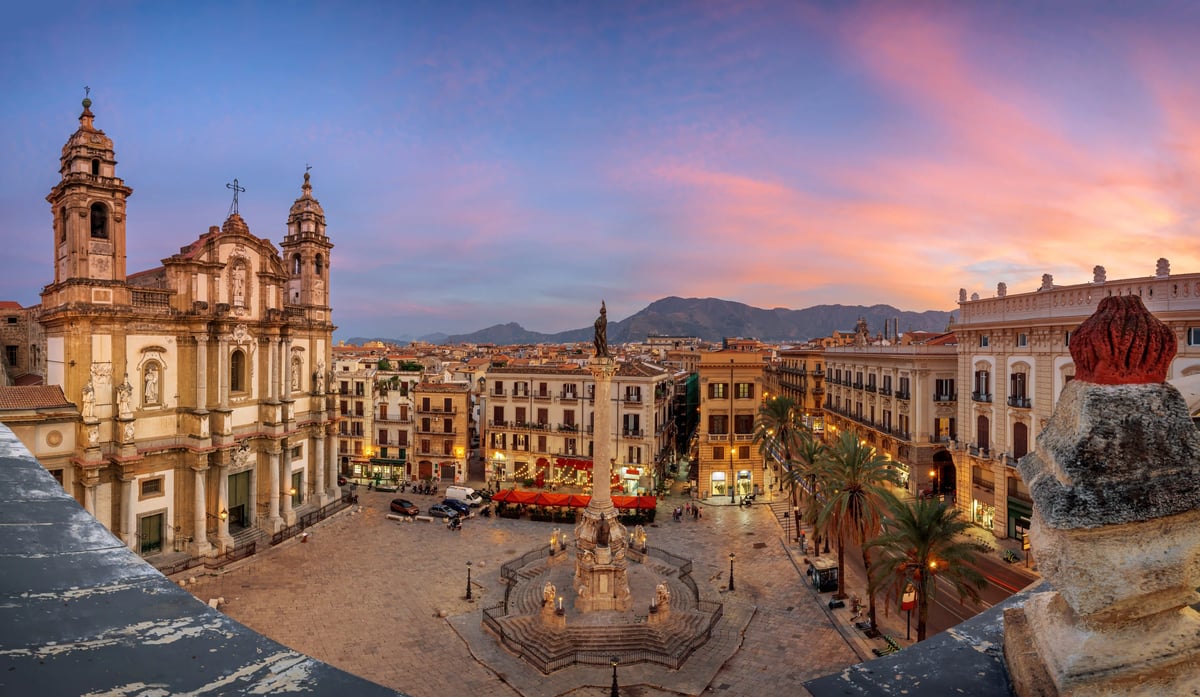 Panoramablick bei Sonnenuntergang auf die Piazza San Domenico in Palermo mit der barocken Kirche, der zentralen Säule und den umliegenden Gebäuden unter einem rosa-orange gefärbten Abendhimmel.