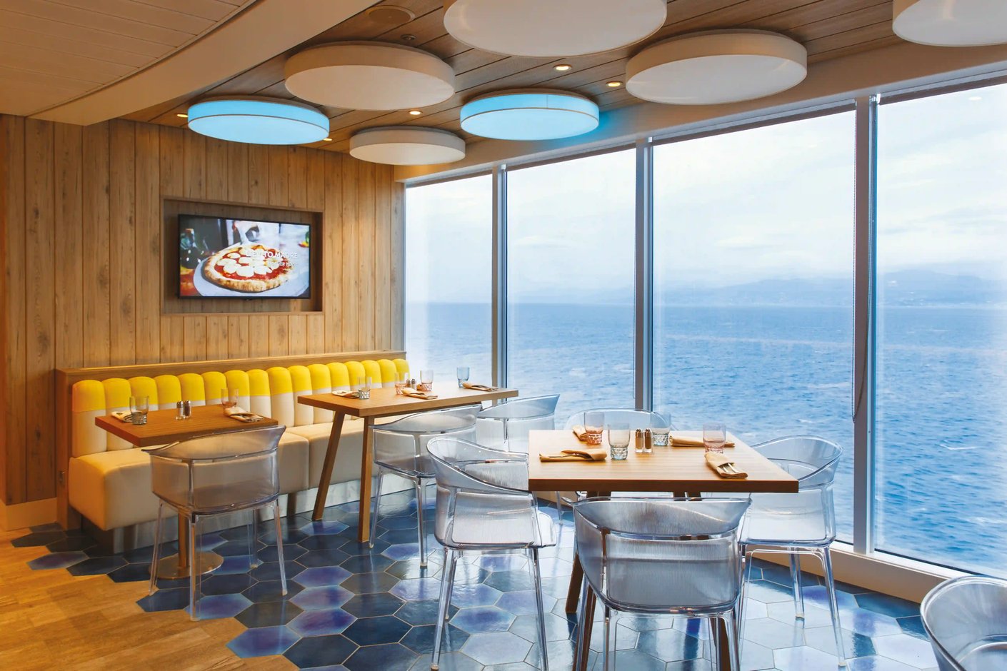 Modernes Schiffsrestaurant mit Holztischen, transparenten Stühlen und einer großen Panoramafensterfront mit Blick auf das Meer