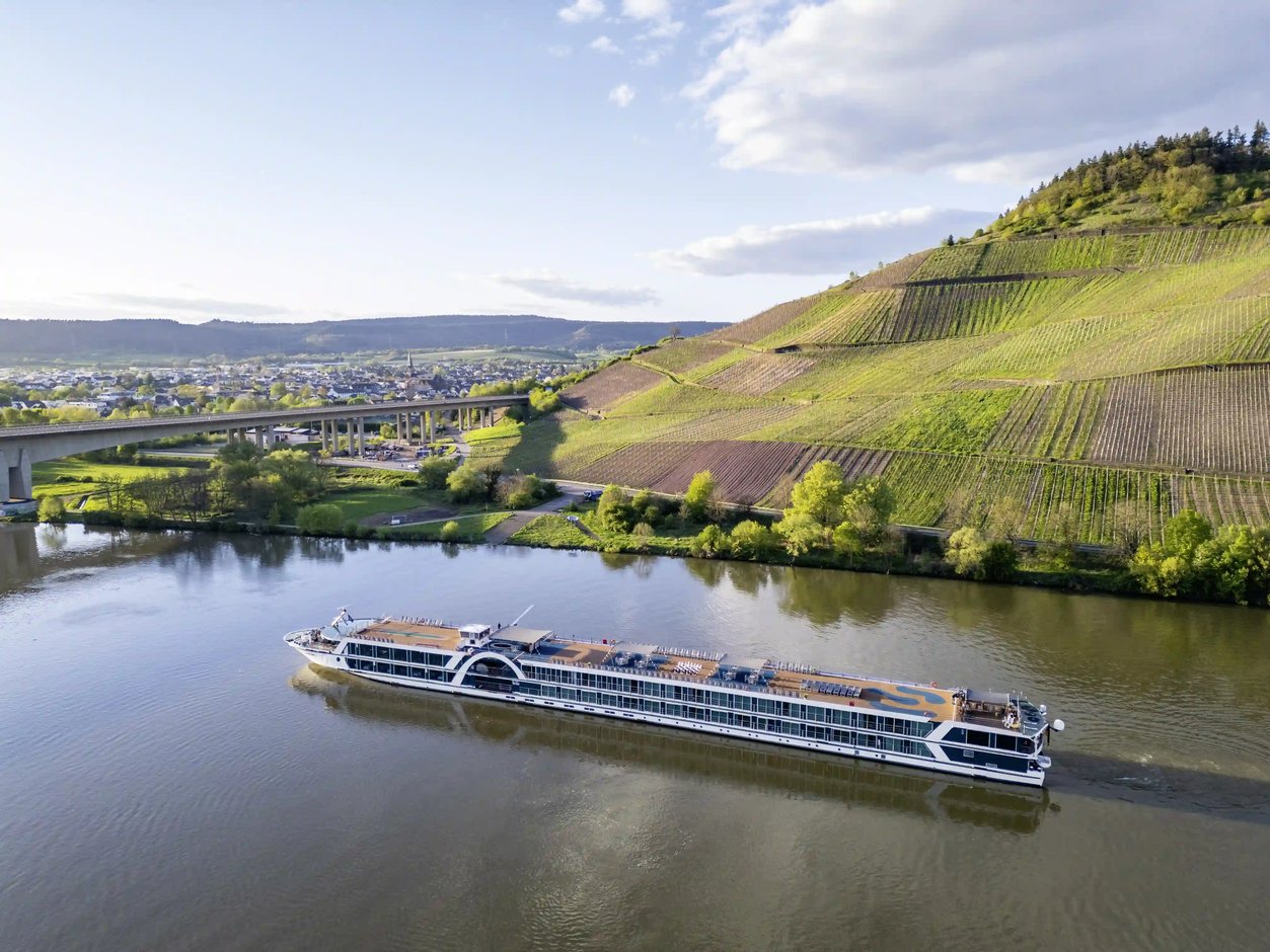 Luftaufnahme des Flusskreuzfahrtschiffs Elegant auf einem breiten Fluss mit Weinbergen, Brücke und Ortschaft im Hintergrund