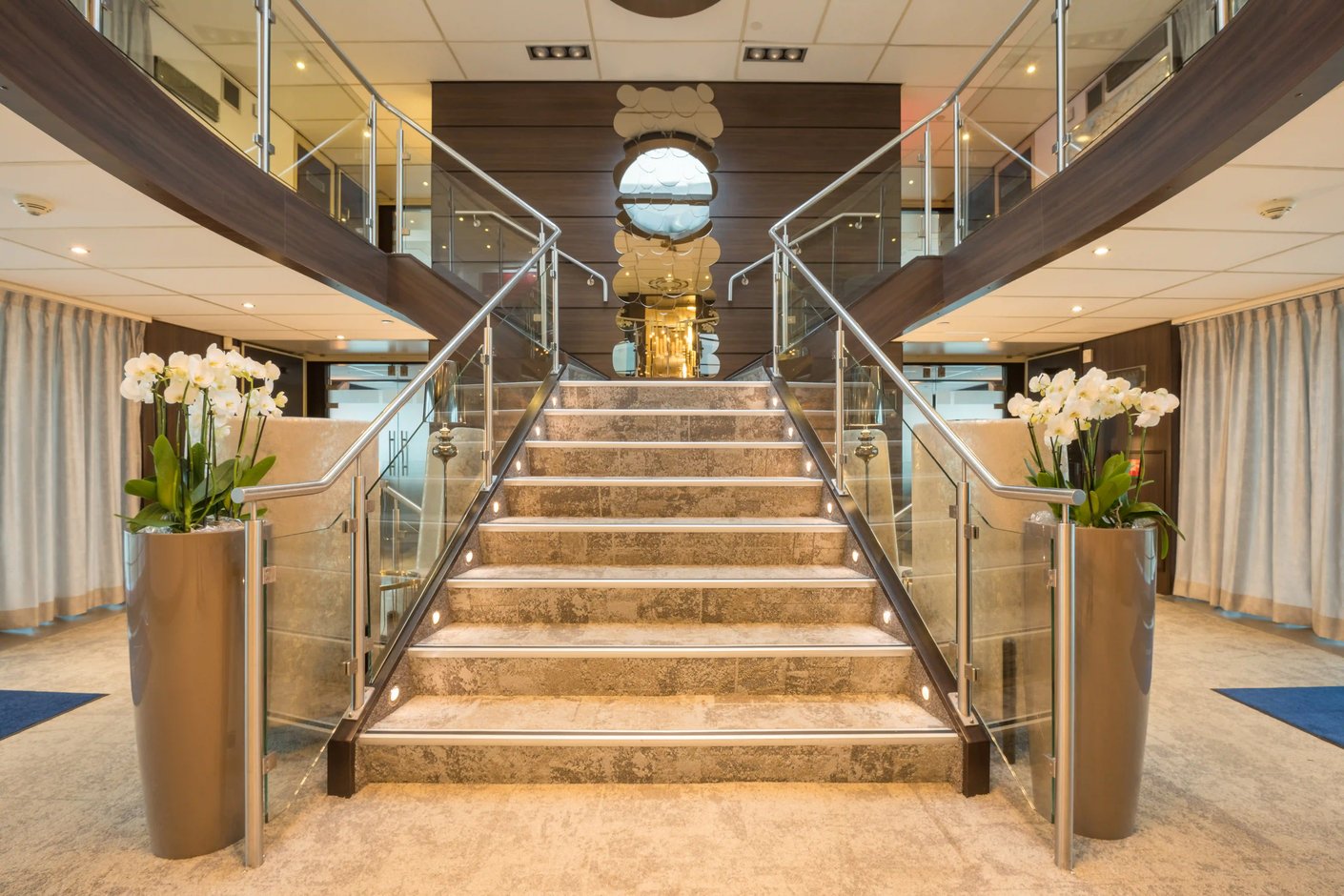 Zentrales Treppenhaus eines Flussschiffes mit Glasgelaendern, symmetrischem Aufbau und moderner Beleuchtung