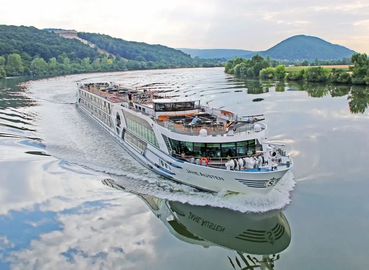 Das weisse Flusskreuzfahrtschiff MS Jane Austen fährt auf einem breiten Fluss vor einer bewaldeten Hügellandschaft