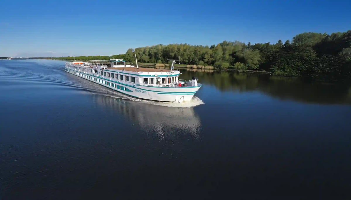 Flusskreuzfahrtschiff faehrt auf ruhigem Gewaesser entlang bewaldeter Uferlandschaft