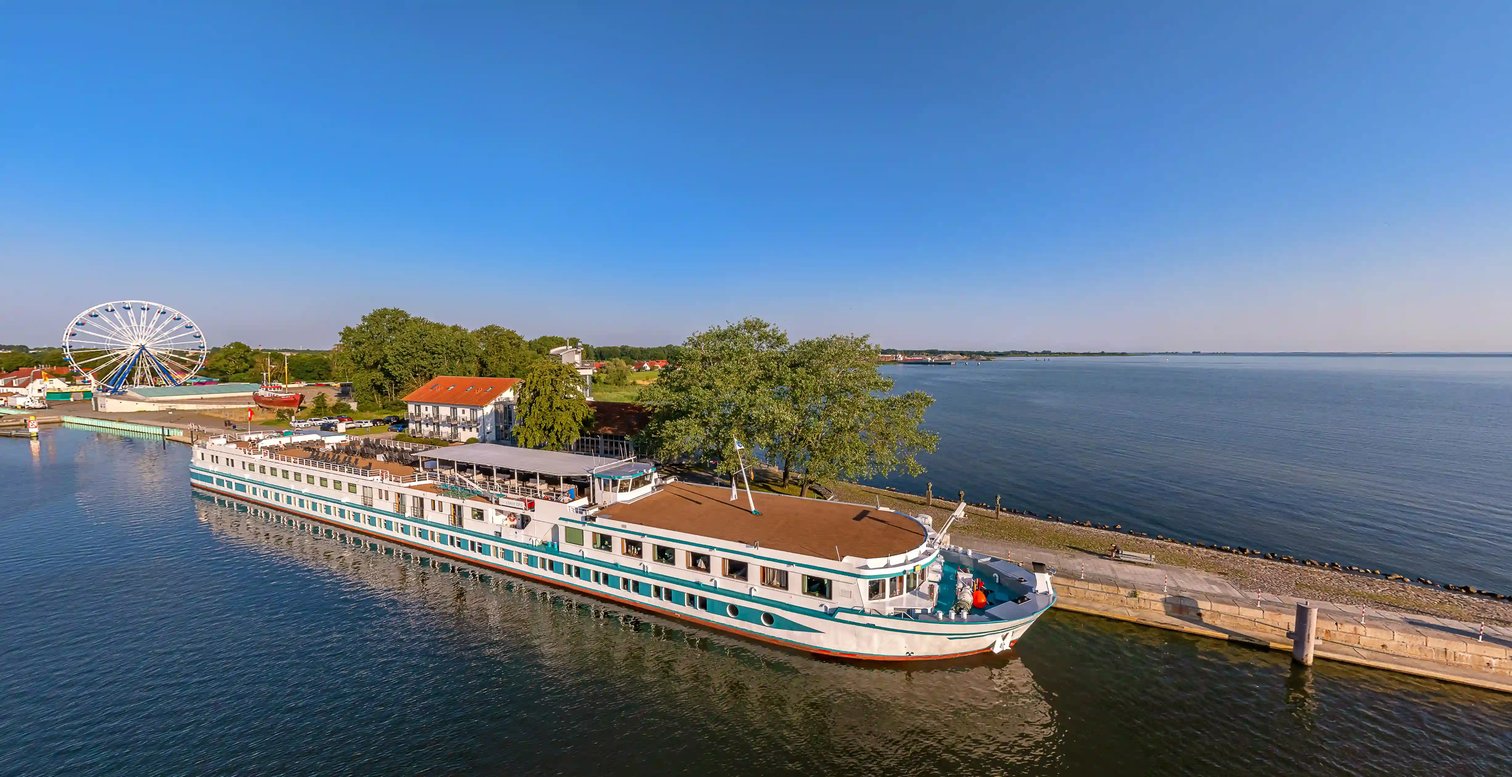 Flusskreuzfahrtschiff am Kai vor einer Küstenlandschaft mit Promenade und Riesenrad