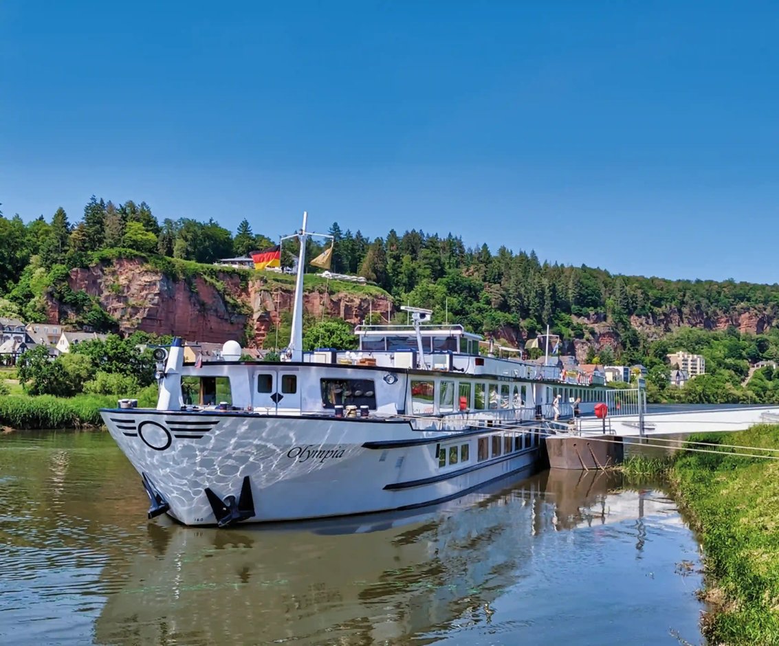 Außenansicht des Flusskreuzfahrtschiffs „MS Olympia“, das an einem Anleger auf einem ruhigen Fluss liegt, umgeben von grünen Hügeln und Felsen bei sonnigem, blauem Himmel