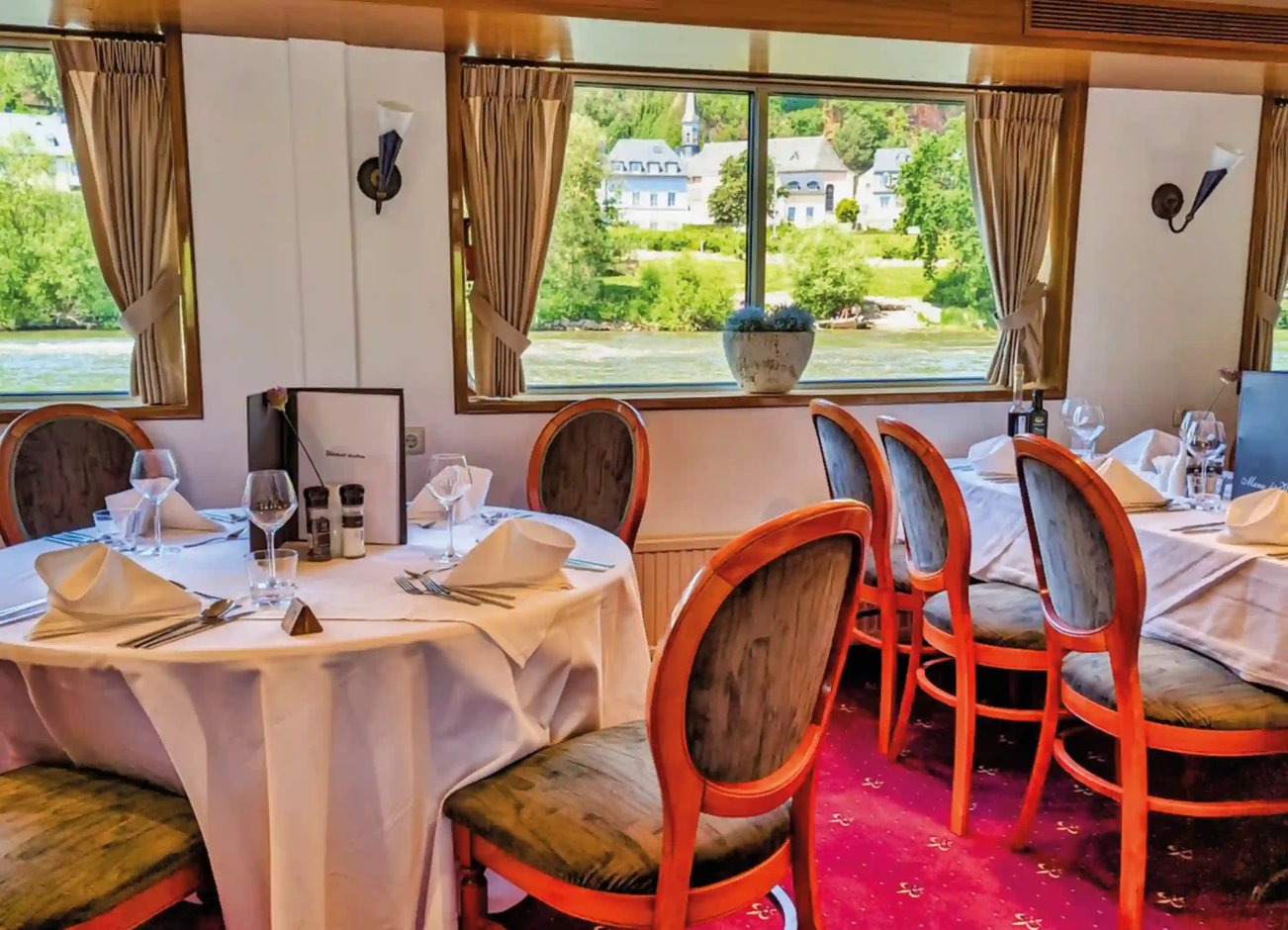 Elegant gedeckter Restaurantbereich an Bord der MS Olympia mit runden Tischen, weißen Tischdecken und großen Panoramafenstern mit Blick auf den Fluss und die Uferlandschaft