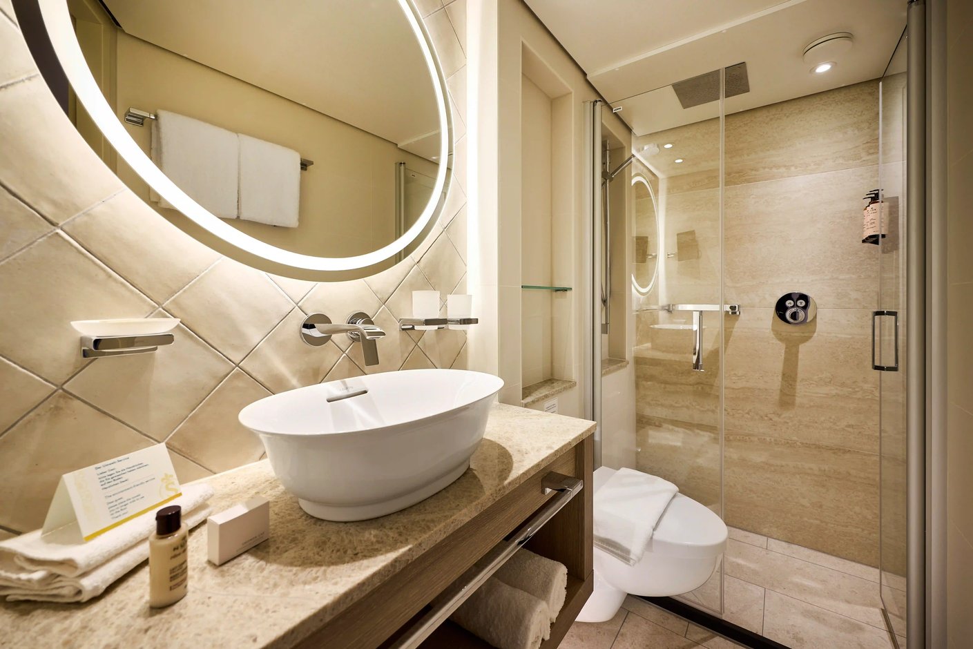 Modernes Badezimmer mit Waschbecken, beleuchtetem Spiegel, Dusche mit Glaswand und hellen Natursteinfliesen.