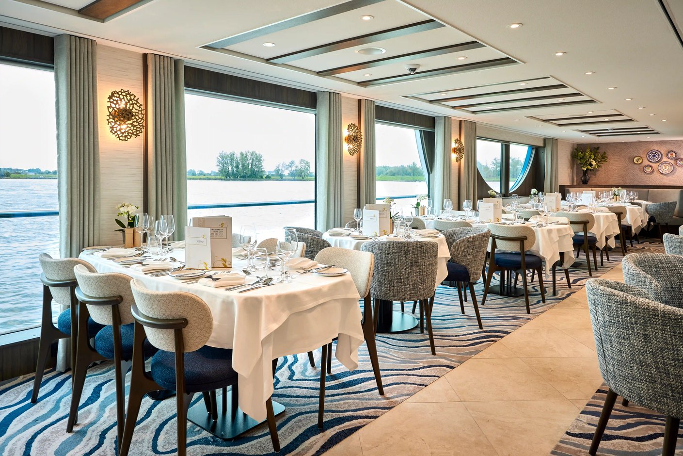 Eleganter Restaurantbereich mit mehreren Tischen, Sesseln und großen Panoramafenstern entlang des Flusses.