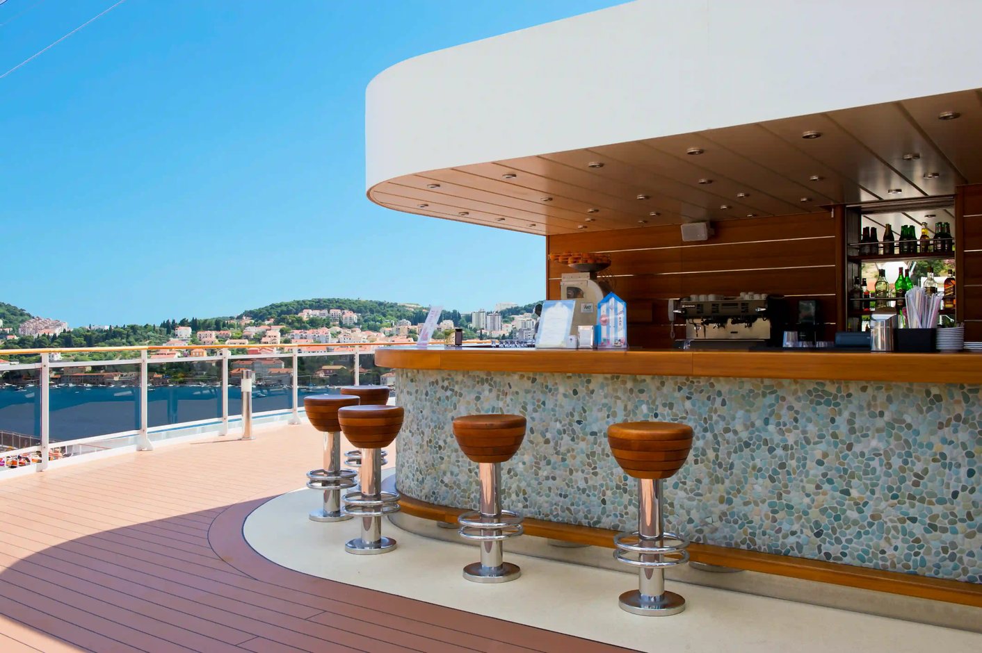 Aussenbar auf der MSC Magnifica mit Bartheke, Barhockern und Blick auf Hafen und Kuestenlandschaft.