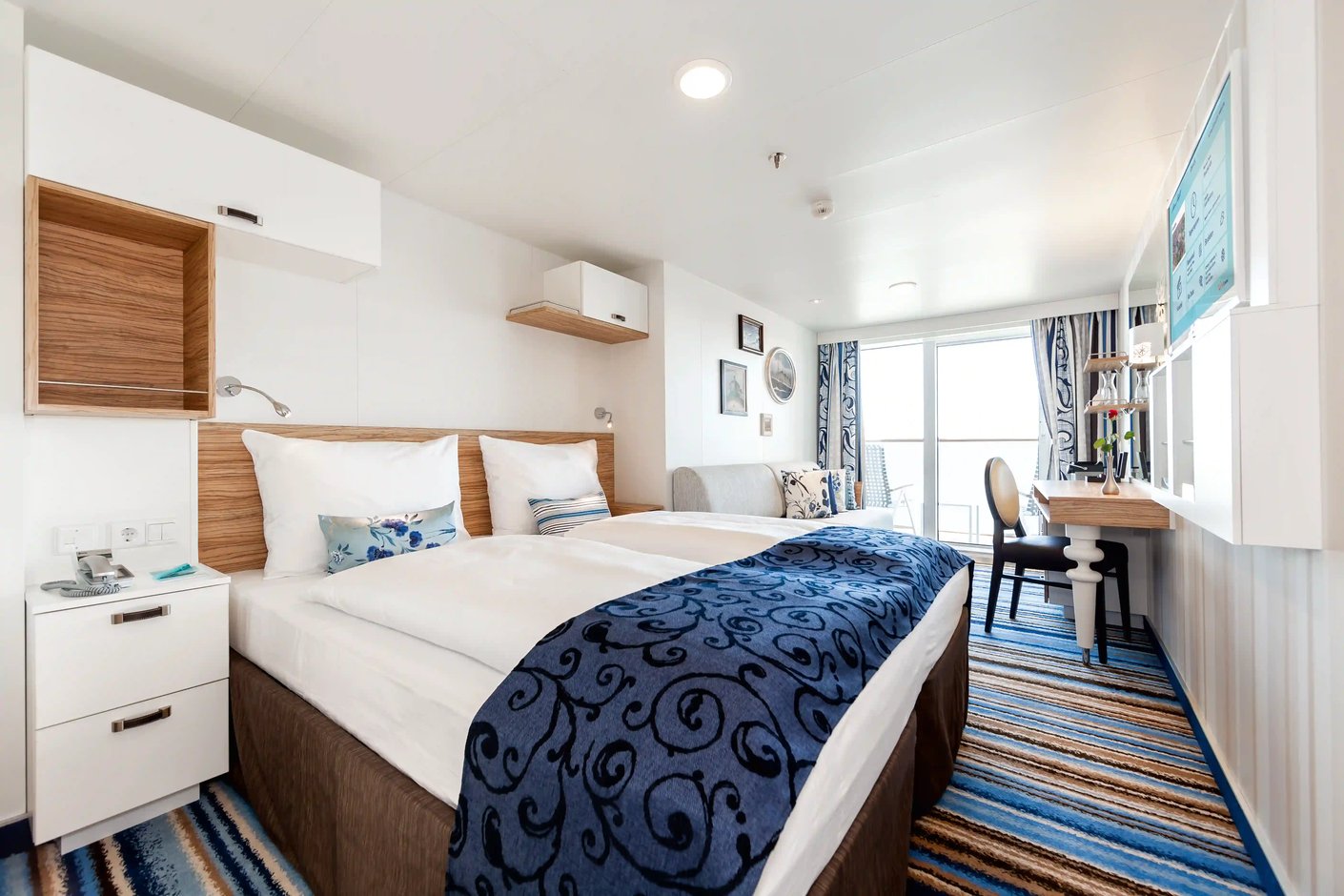 nnenansicht einer Balkonkabine auf der Mein Schiff 3 mit einem Doppelbett, einem kleinen Sofa, einem Schreibtisch und einer großen Balkontür mit Meerblick.