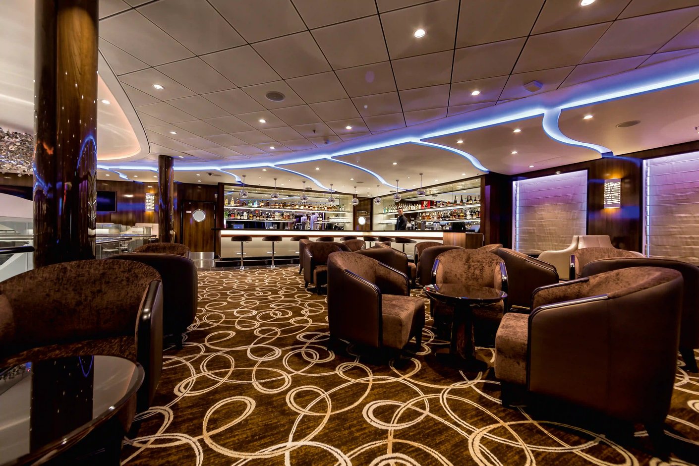 Elegante Bar an Bord der Mein Schiff 3 mit gedimmter Beleuchtung, bequemen braunen Sesseln, einem gemusterten Teppich und einem gut ausgestatteten Bar-Tresen im Hintergrund