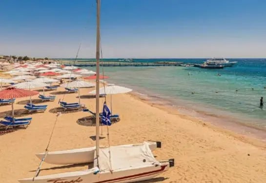 Sandstrand am Roten Meer mit Liegestühlen, Sonnenschirmen und Segelboot vor dem Hotel.