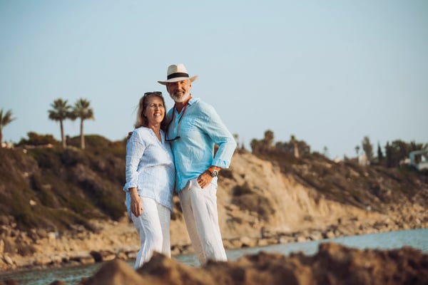 Älteres Paar steht lächelnd am Meer vor felsiger Küste bei sonnigem Wetter.