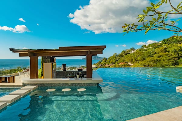 Moderne Poolbar in einem Infinity Pool mit Blick auf das Meer und grüne Hügel bei sonnigem Wetter