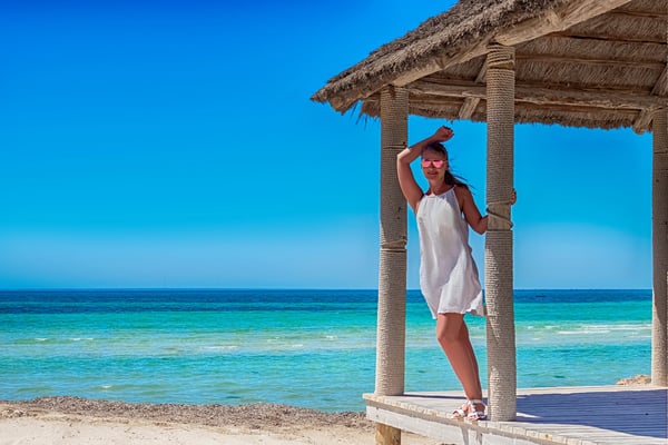 Frau im Sommerkleid steht unter einer Strandhütte am türkisblauen Meer.
