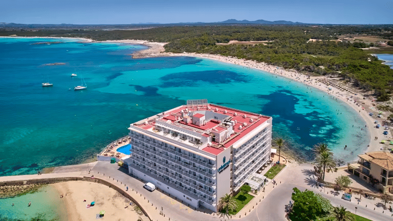 Modernes Hotelgebäude an einer Bucht mit Sandstrand, türkisblauem Meer, Küstenstrasse und Naturgebiet im Hintergrund.