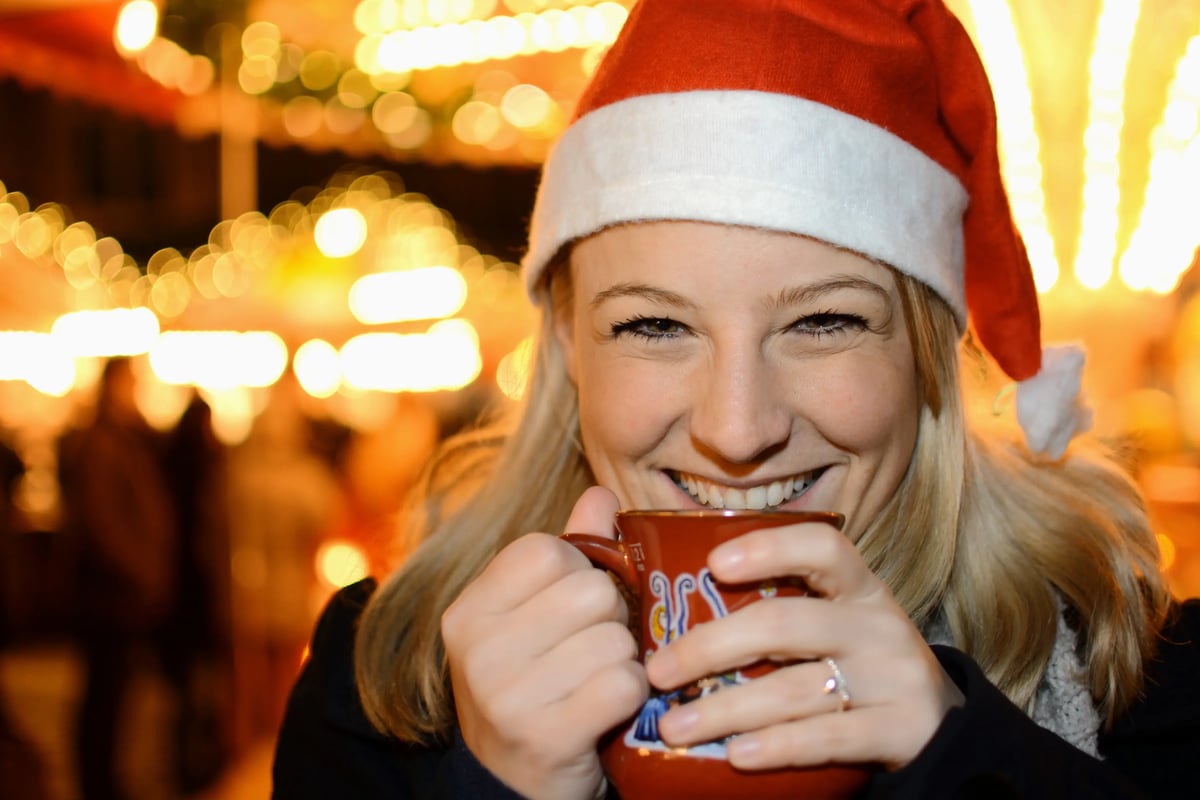 Lächelnde Frau mit Weihnachtsmütze trinkt ein heisses Getränk auf einem Weihnachtsmarkt.