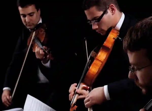 Drei Musiker in schwarzen Anzügen spielen Geige in einem Konzert, ein Notenblatt liegt im Vordergrund.