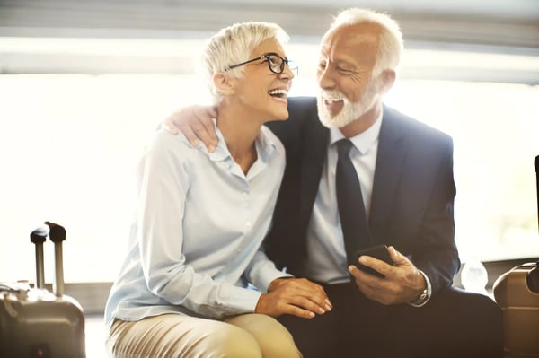 Älteres Paar sitzt mit Koffern in einem hellen Wartebereich, lacht und schaut gemeinsam auf ein Smartphone.