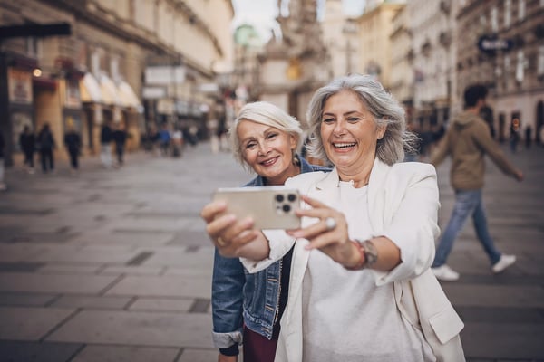 Zwei ältere Freundinnen machen lachend ein Selfie in einer belebten Altstadt