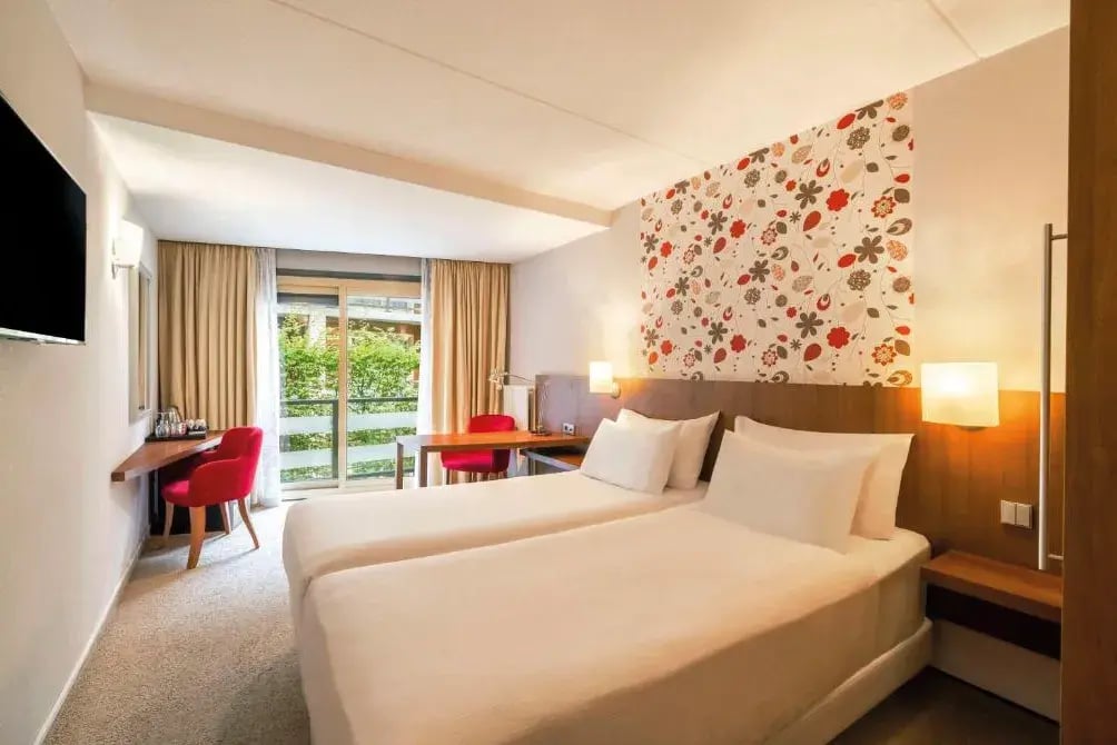Chambre d’hôtel lumineuse avec deux lits, bureau, chaise rouge et grande fenêtre donnant sur la verdure