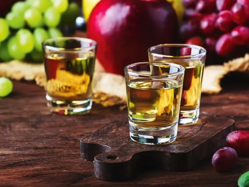 Trois verres de rakija traditionnelle devant des fruits frais sur une table rustique en bois