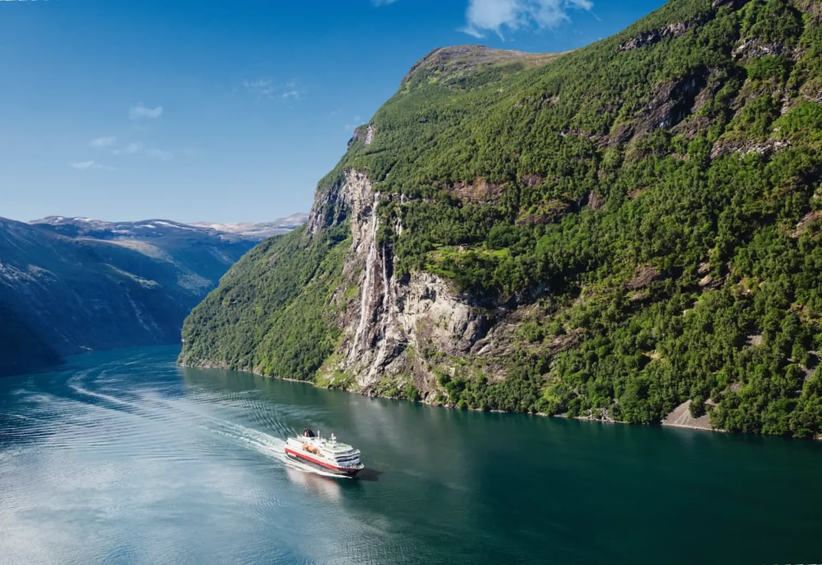 Hurtigruten-Schiff fährt durch einen norwegischen Fjord mit steilen Felswänden und Wasserfall.