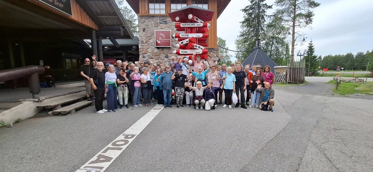 Reisegruppe posiert am Polarkreis vor einem roten Wegweiser in Rovaniemi