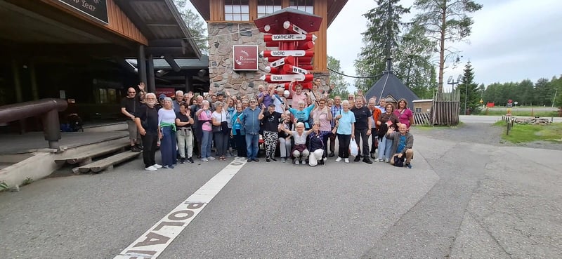 Reisegruppe posiert am Polarkreis vor einem roten Wegweiser in Rovaniemi
