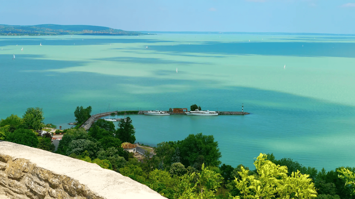 Panoramablick über den türkis schimmernden Plattensee mit Segelbooten und einer Anlegestelle.