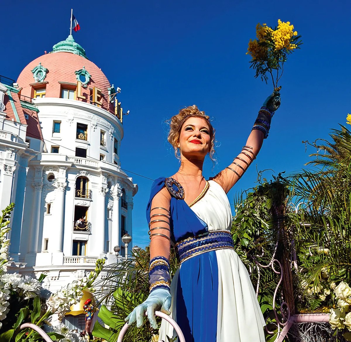 Femme en costume historique tenant un bouquet de mimosa devant un char du Carnaval de Nice.