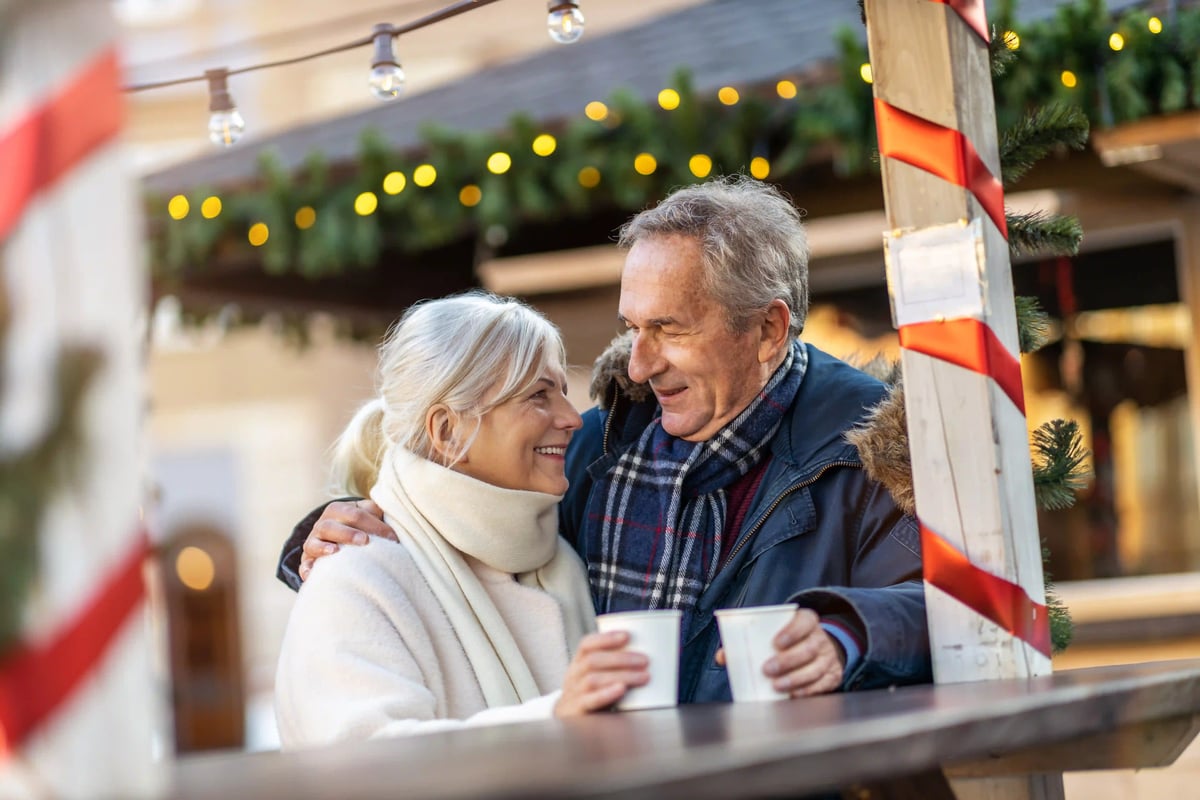 Älteres Paar auf einem Weihnachtsmarkt trinkt gemeinsam ein heisses Getränk und lächelt sich an. ALT-Text Französisch: